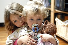 To børn. Det yngste barn har en sut i munden. Det ældste barn krammer det yngste bagfra.